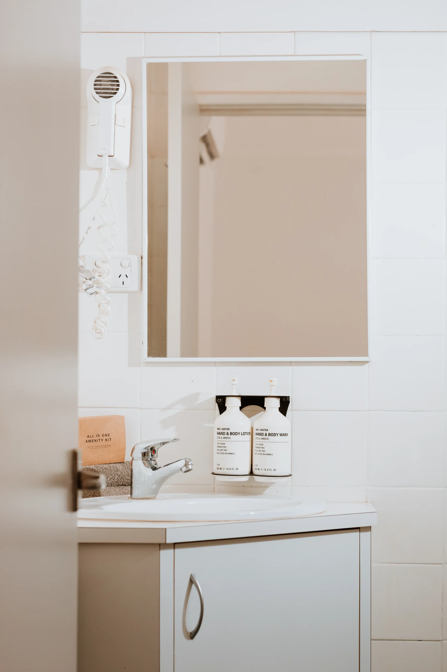Photo of bathroom with corner vanity unit including: hairdryer, Ink & Water handwash, towels and a mirror