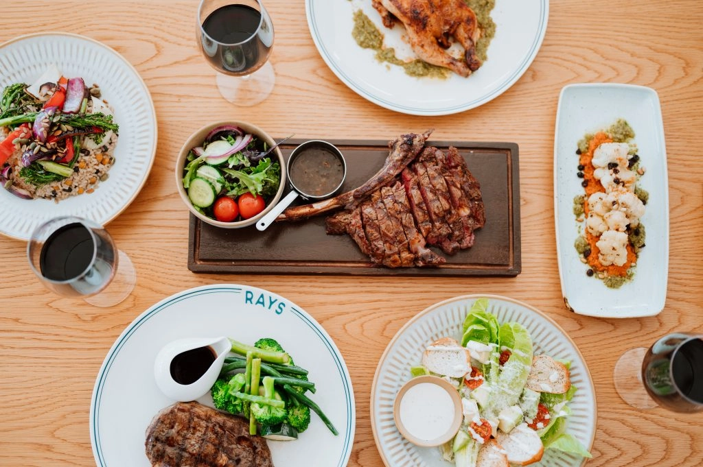 A spread of food including steaks, cooked vegetables, salads, and red wine at a pub